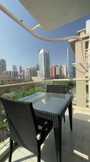 Balcony terrace with glass table and two wicker chairs in the foreground, overlooking Dubai high-rise skyline and clear blue sky.