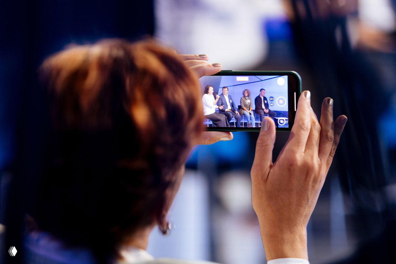 Кадр с конференции: зритель снимает сцену на смартфон, видна панель спикеров и активная запись мероприятия для социальных сетей.