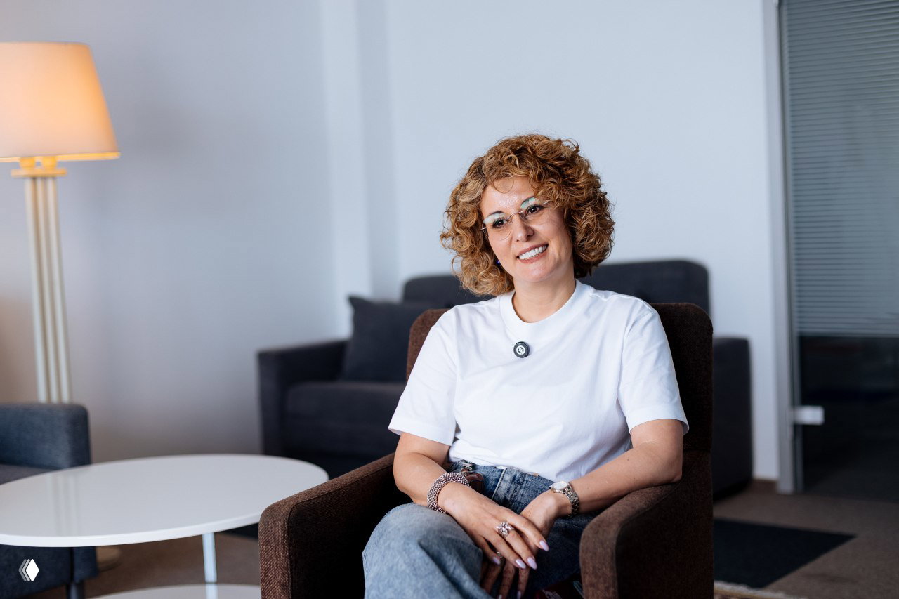 Портрет Ангелины Самадовой в неформальной обстановке: улыбающаяся ведущая сидит в кресле, интерьер зоны отдыха школы управления.