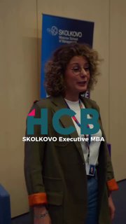 Женщина в деловом костюме выступает на фоне баннера SKOLKOVO и логотипа HCB; вертикальный кадр, конференционная обстановка, живой ракурс.