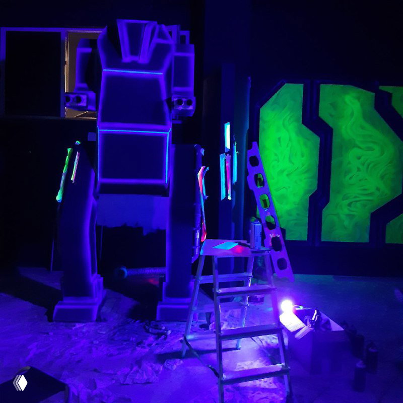 Ночная сцена арены с ультрафиолетовыми лампами: флуоресцентные рисунки, высокий роботоподобный силуэт и стремянка в зоне работ.