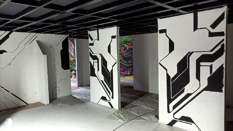 Ряд вертикальных панелей с завершёнными чёрными узорами на белом фоне, тёмный потолок и разноцветные граффити на дальней стене.