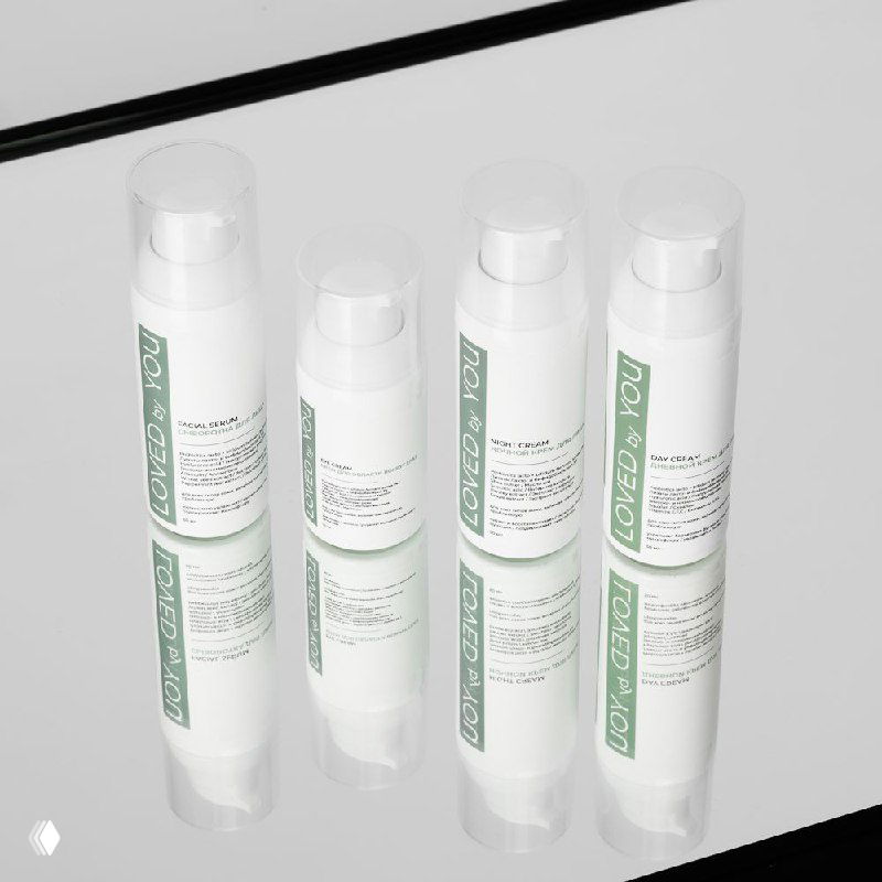 Несколько флаконов средств Loved by You на зеркальной поверхности с отражением; белая упаковка с зелёной этикеткой и аккуратным брендингом.