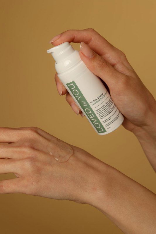 Крупный план рук наносит средство из флакона Loved by You: видно дозатор и текстуру средства на коже на бежевом фоне.