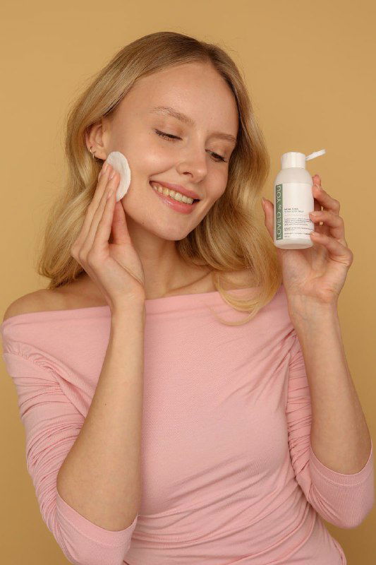 Женщина наносит тоник Loved by You на ватный диск, держит флакон и улыбается — демонстрация способа применения продукта.