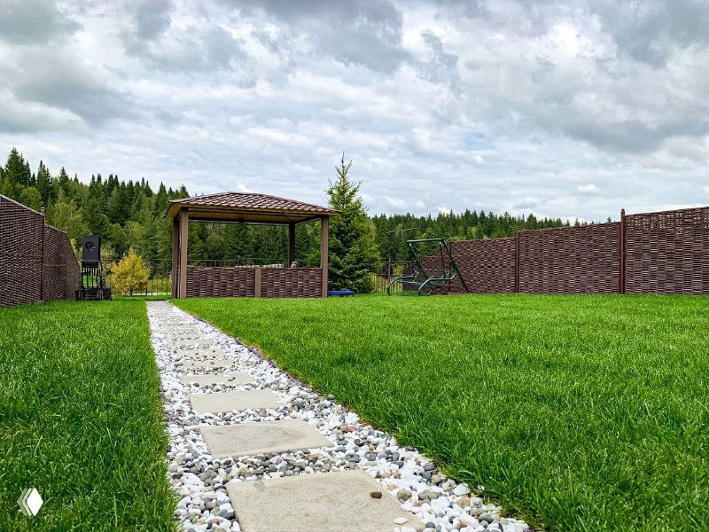 Фокус на аккуратный газон и центральную дорожку из плит; видна беседка, ограждение участка и лесной массив под сероватым небом.