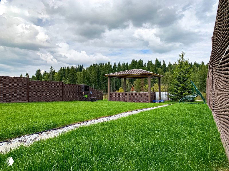 Общий вид участка с густым рулонным газоном, деревянной беседкой и забором по периметру; на заднем плане — хвойный лес и пасмурное небо.