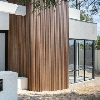 Угловой фрагмент фасада дома, покрытый вертикальными панелями из ДПК, крупный план показывает фактуру, стыки и цвет покрытия на фоне окна и земли.