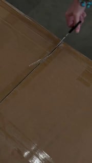 Кадр из видео распаковки: рука с канцелярским ножом разрезает картонную коробку с доставкой комплекта мебели, момент распаковки