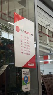 Стеклянная табличка с логотипом LETOMARKET и расписанием работы магазина, размещённая внутри торгового зала рядом с витриной.