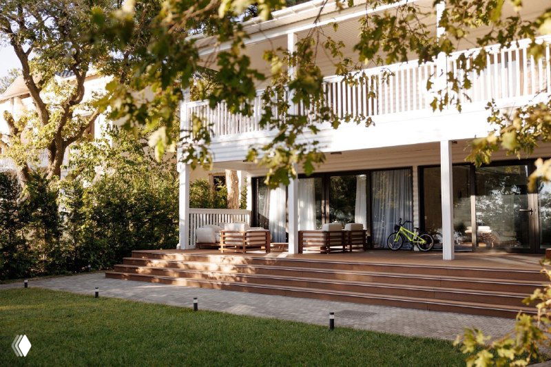 Терраса с широким настилом из ДПК перед современным домом, мебель для отдыха размещена на веранде, зелёная зона вокруг.