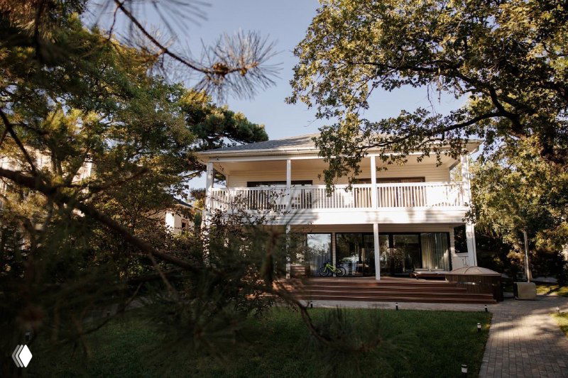 Фасад загородного дома с большой террасой и настилом из композитной древесины (ДПК), окружённый деревьями и газоном.