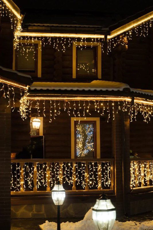 Деревянный дом вечером, украшенный свисающими светодиодными гирляндами-водопадом вдоль балкона и крыши; тёплое декоративное освещение на фасаде.