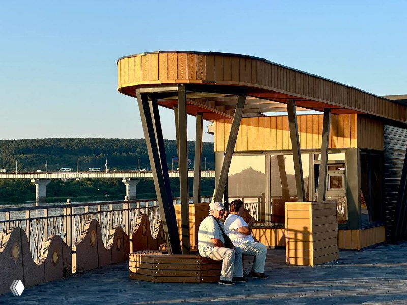 Площадка на набережной: люди на лавочках у павильона с тёплым оттенком ДПК, металлические опоры и берег реки на фоне при дневном свете.