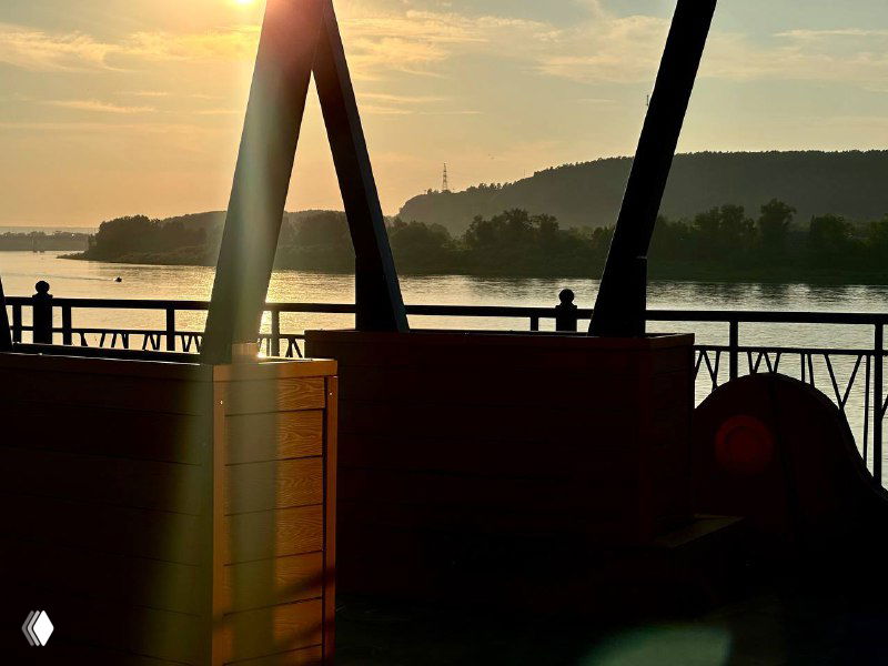 Силуэт опор и посадочных мест на фоне заката: фактура ДПК вблизи, отражение солнца на реке и прогулочная зона на набережной.