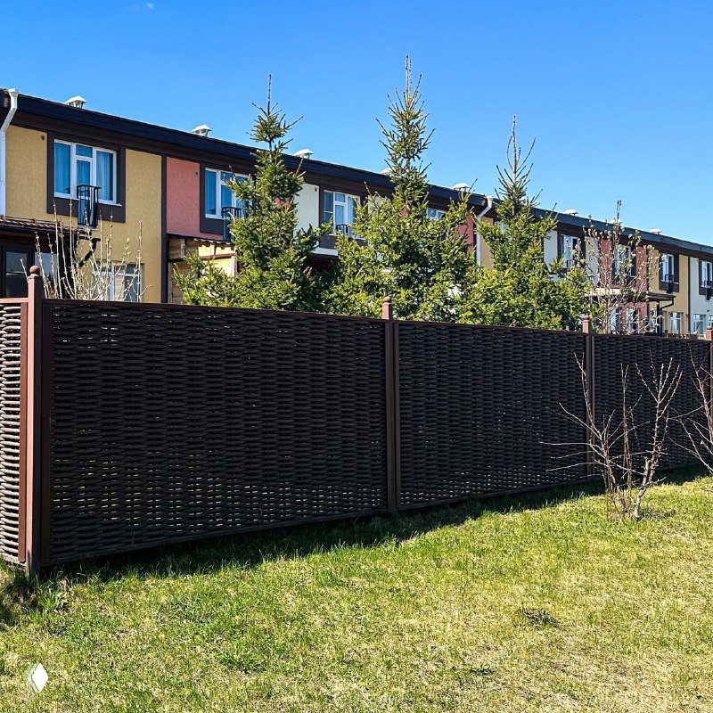Длинная секция забора из полимерной лозы вдоль газона и ряд таунхаусов на заднем плане; наглядный пример зонирования участка и защиты от взглядов.