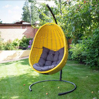 Жёлтое плетёное кресло‑кокон на подставке на зелёном газоне, вокруг деревья и садовый забор, наружная установка в частном дворе.