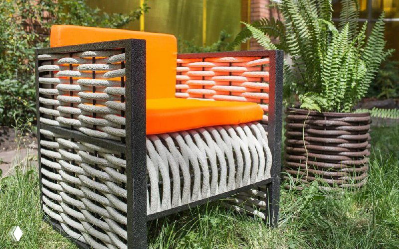 Садовое кресло с плетёной спинкой и яркой подушкой рядом с горшками и листвой — пример мебели из полимерной лозы на открытой веранде.