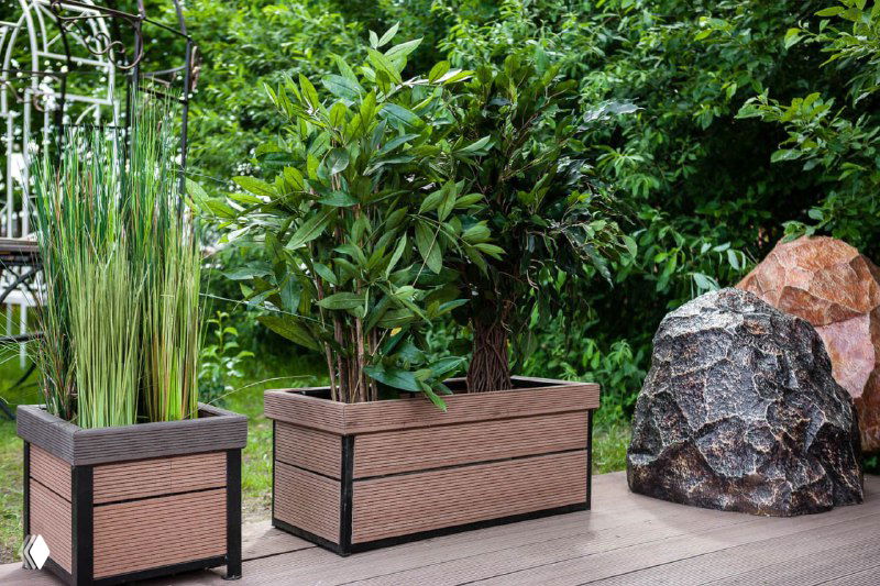 Два декоративных кашпо из светло‑коричневого ДПК на террасе среди зелёных растений — аккуратное оформление цветочных композиций.