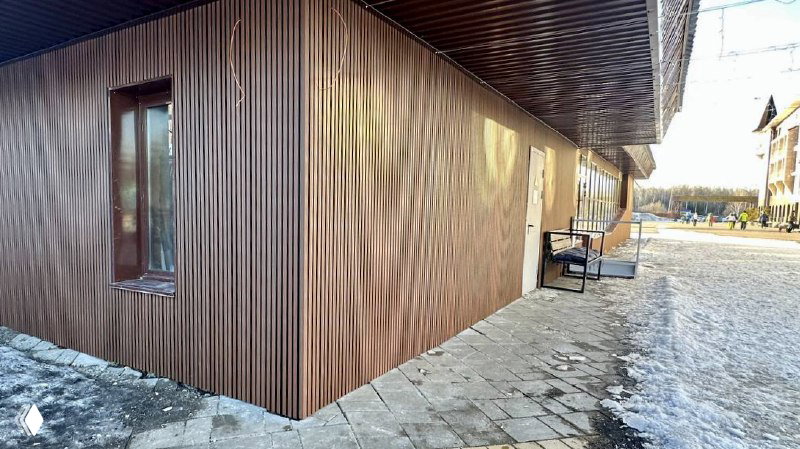 Вид вдоль стены ресторана «Танай» с вертикальными брусками ДПК, показан монтаж панелей, тротуар и снежные участки в окружении зданий.