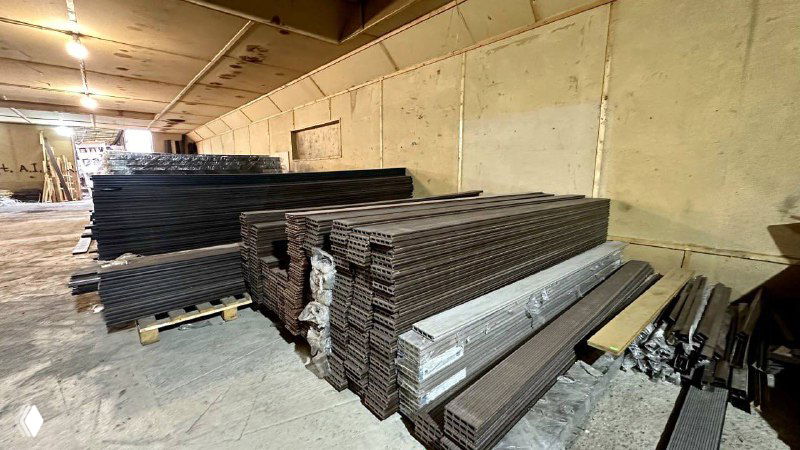 Общий вид склада с уложенными пачками заборной и террасной доски, видны паллеты, упаковка и промежутки между стопками