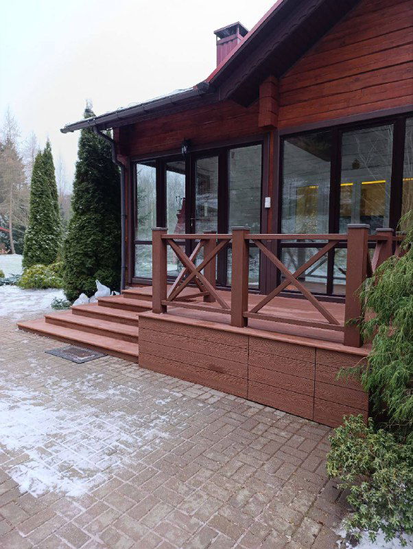 Ещё один уголок красной террасы с лестницей и площадкой у входа, видно сочетание доски и облицовки фасада дома в естественном освещении.