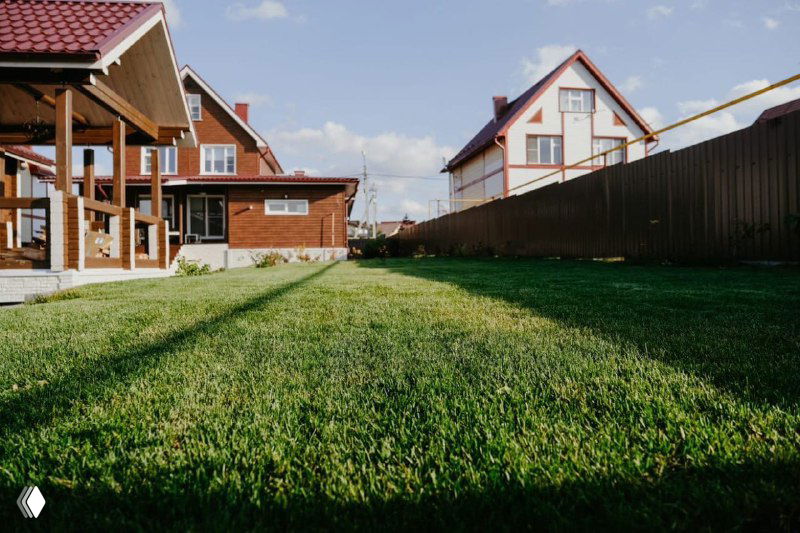 Продольный вид ухоженного газона между домами и забором, демонстрация ровного роста и результата укладки рулонного покрытия.