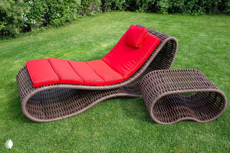 Шезлонг из полимерной лозы тёмно-коричневого цвета на зелёной лужайке; волнообразная форма и яркая красная подушка выделяются на фоне травы.