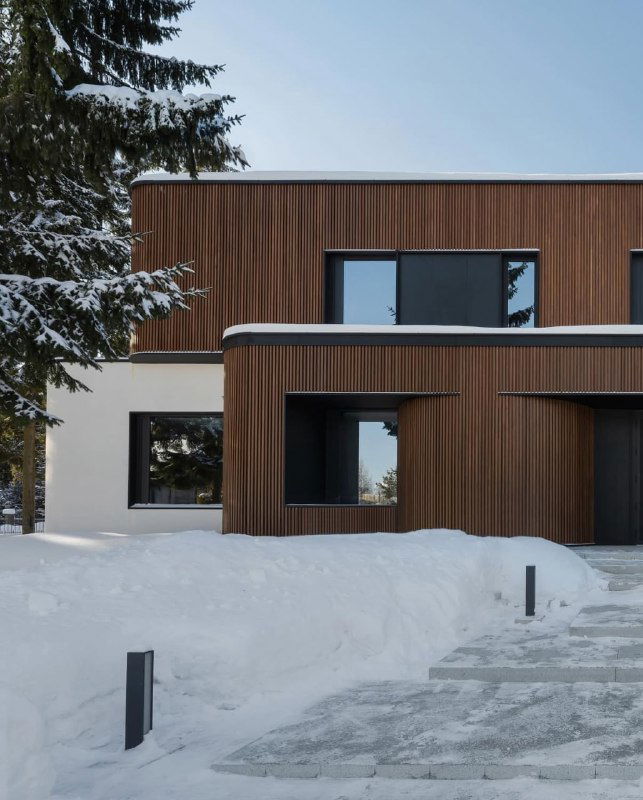 Фасад дома зимой: брусковые панели ДПК частично покрыты снегом, аккуратная тропинка и хвойные деревья создают атмосферу холодного сезона.