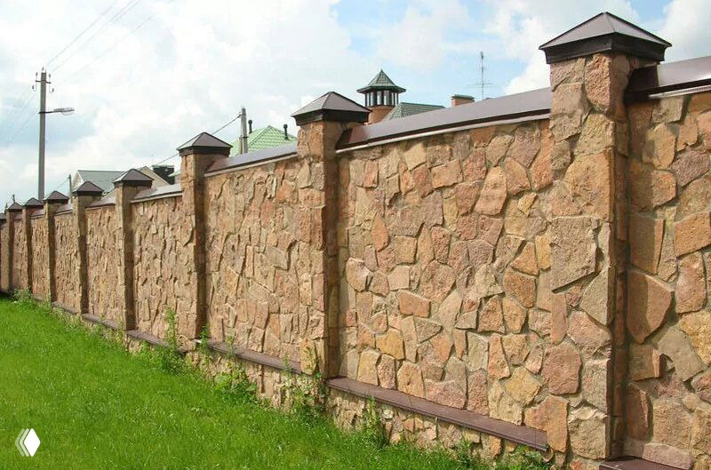 Высокий забор из натурального камня и кирпича с колоннами на фоне зелёной лужайки; показывает массивность и долговечность каменных ограждений.