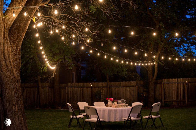 Подвешенные фонарики и огоньки в саду над столом создают тёплую атмосферную подсветку для вечерних встреч на участке.