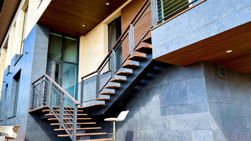 Угол дома с открытой металлической лестницей и террасой из ДПК, сочетание фасадных материалов и ограждения балкона.