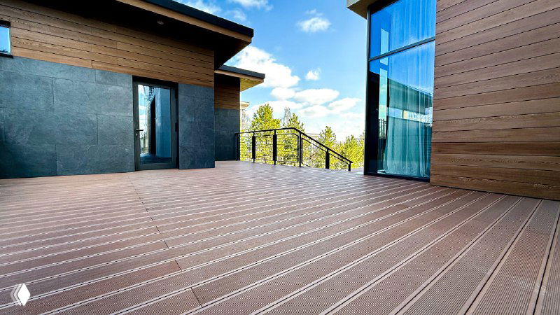 Панорамный вид террасы из ДПК ближе к входу, демонстрация цвета 'песок' и сочетания с тёмными фасадными панелями дома.