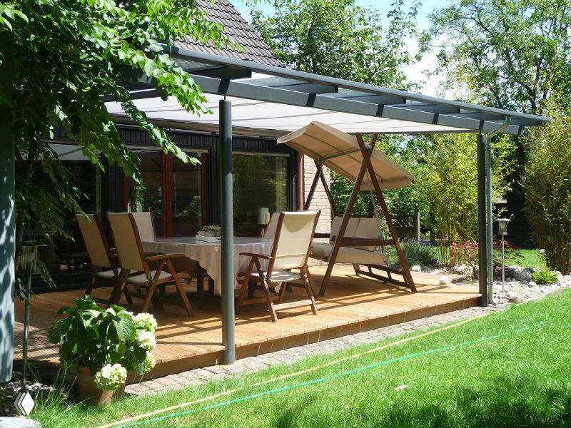 Современная терраса с навесом из прозрачного поликарбоната, столовой зоной и шезлонгами на фоне зелёного сада и газона.