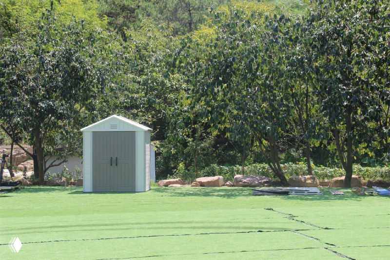 Одиночный компактный пластиковый хозблок на зелёной площадке у дерева, подчёркивает размеры модели для небольшого участка.