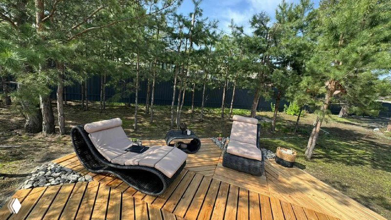 Открытая терраса: две шезлонговые лежанки и дизайнерское кресло из полимерной лозы на деревянном настиле среди зелени и сосен.