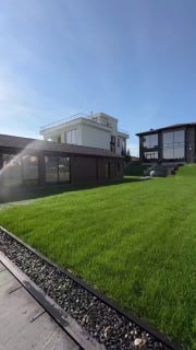 Ровный зелёный рулонный газон на участке перед современным домом, солнечная погода, аккуратная дорожка и система полива на заднем плане.