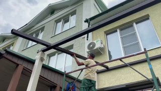 Мастер устанавливает металлический каркас террасы у фасада двухэтажного дома, видны рабочие, лестница и опоры монтажных элементов