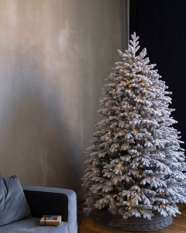 Высокая пушистая заснеженная искусственная ёлка в углу комнаты рядом с креслом — минималистичный праздничный интерьер.