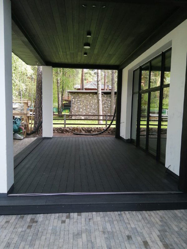 Просторная тёмная терраса под навесом с однотонной доской POLIVAN SINGARAJA, вид с уровня пола на покрытие и дверь в дом.