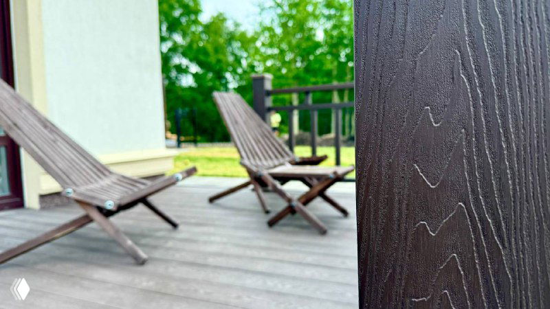 Крупный план текстуры и оттенка коричневого настила из композитной доски Polivan SINGARAJA, стоящие складные кресла и зелёный фон участка на заднем плане.