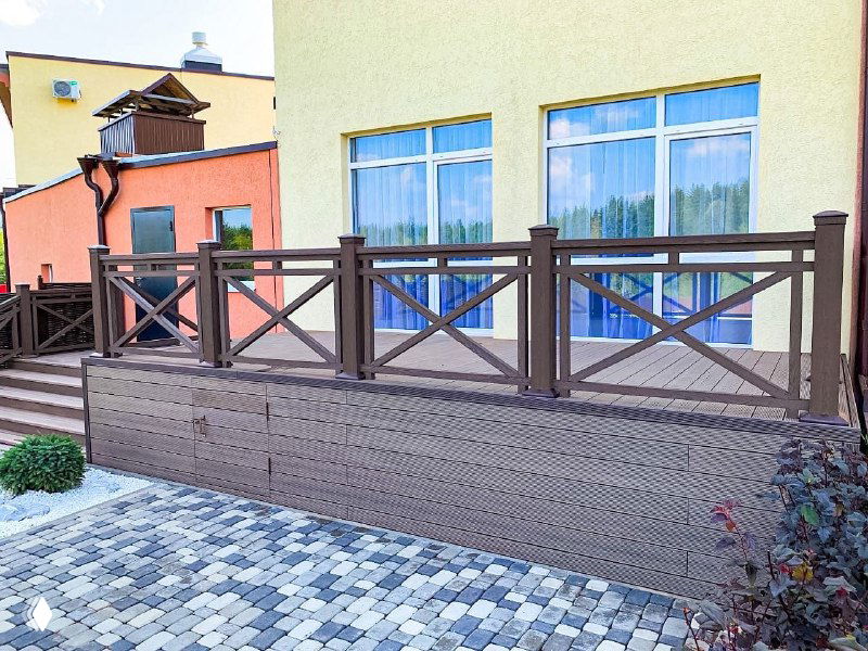 Крупный план ограждения и цоколя террасы из композитной доски, видно текстуру ДПК и сочетание с серой брусчаткой у дома.