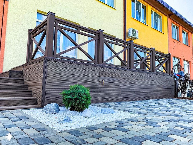 Угол обзора на террасу с лестницей и цоколем из композитной доски, небольшой декоративный куст и аккуратный каменный отмосточный пятачок.