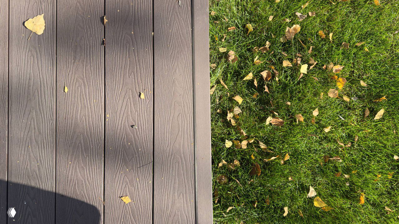 Крупный план настила из ДПК рядом с зелёной лужайкой: видна текстура досок, осенние листья и контраст покрытия и травы.