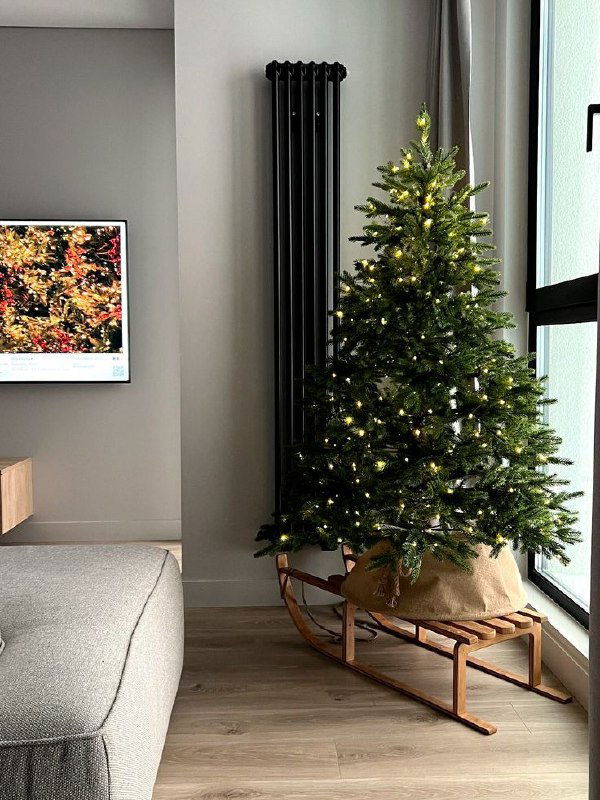 Средняя по размеру ёлка у панорамного окна и чёрного радиатора, украшенная тёплыми лампочками; современная гостиная и зона отдыха на фоне.