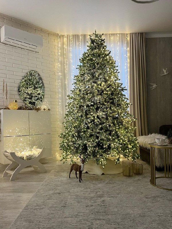 Высокая пушистая ёлка с множеством тёплых огней в просторной светлой гостиной с белыми стенами, ковром и праздничными украшениями на полу.