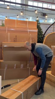 Менеджер распаковывает коробку с искусственной ёлкой в торговом пространстве, вокруг видна мебель и упаковочные материалы.