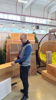 Мужчина раскрывает упаковку с ёлкой на полу торгового зала; вокруг коробки и часть выставочного пространства магазина видны ясно.