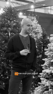 Чёрно-белая фотография в магазине: мужчина с бокалом стоит между несколькими искусственными ёлками, демонстрация ассортимента LetoMarket.