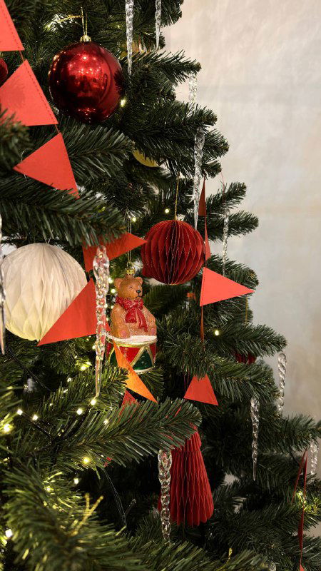 Близкий план веток ёлки с красными кисточками, шариками и декоративными элементами; акцент на текстуре ветвей и мерцающих огоньков праздничной конструкции.